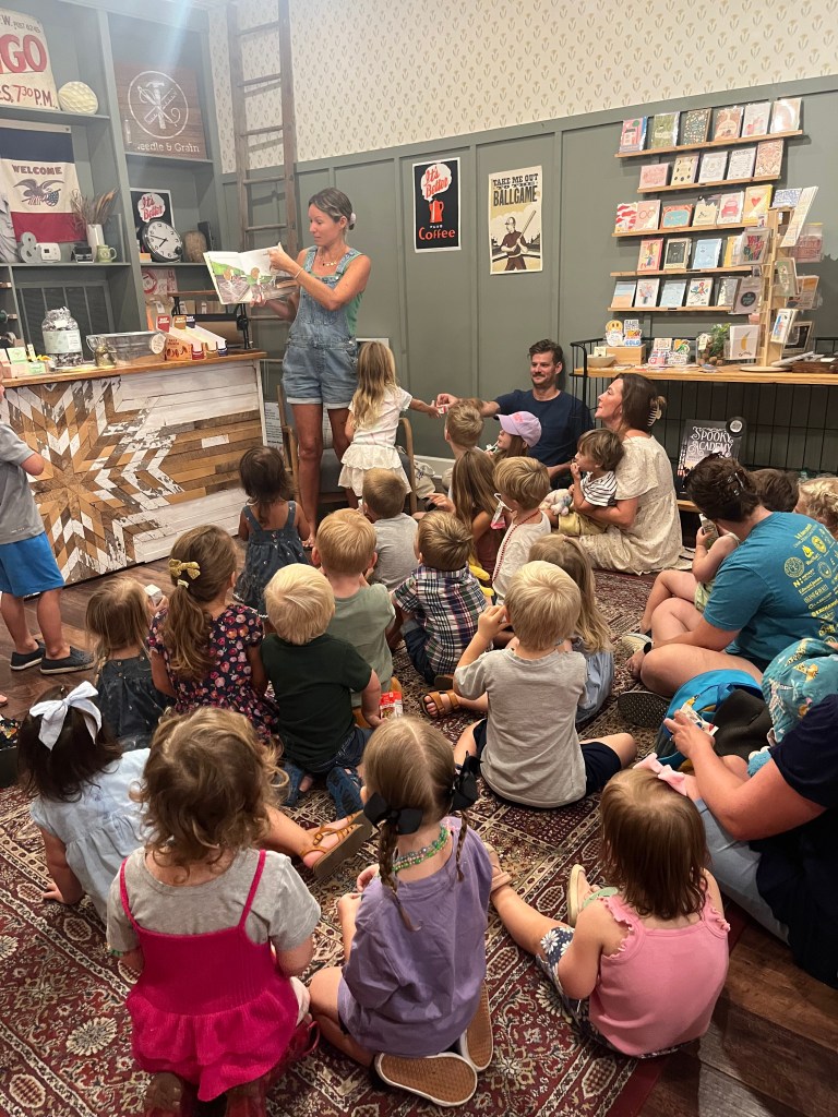 Children sitting on a carpet during a Needle and Grain story time session while Bradley Mountain staff read a book.