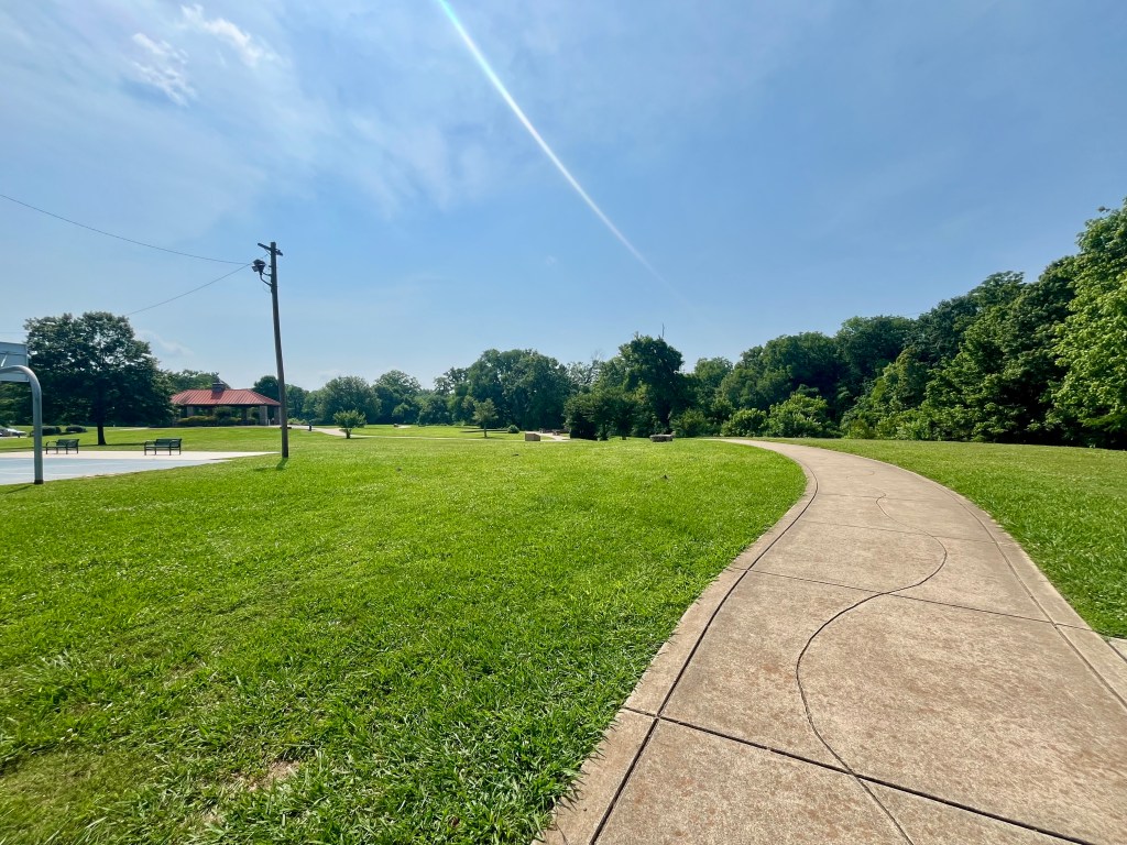 Paved stroller-friendly trail at Riverwalk Park in Columbia Tennessee with trees, a basketball court, and a pavilion in the distance.