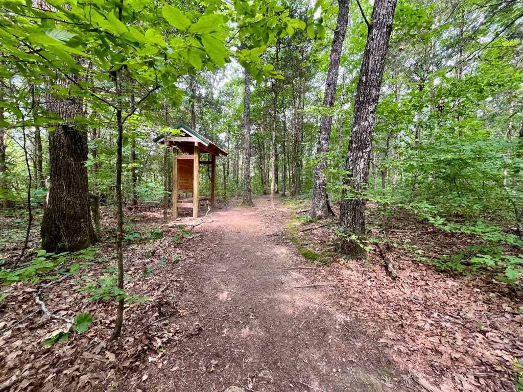 Entrance to the Cheeks Bend Bluff View Trail at the Duck River Complex State Natural Area in Columbia Tennessee with a wooden trailhead kiosk on the left surrounded by trees.