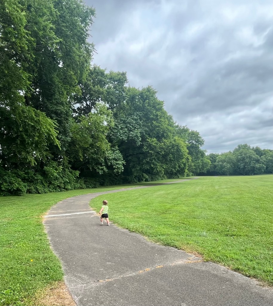 Child running along the paved walking trail at Williams Spring Park in Mount Pleasant Tennessee, with trees on one side and an open grassy field on the other.